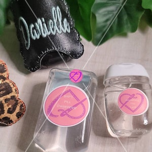 Może przedstawiać: Dwie przezroczyste plastikowe butelki z płynem do dezynfekcji rąk z r&oacute;żowymi i białymi etykietami z napisem "Stitching the Love". Większa butelka ma czarną sk&oacute;rzaną obudowę z wyhaftowanym imieniem "Danielle".