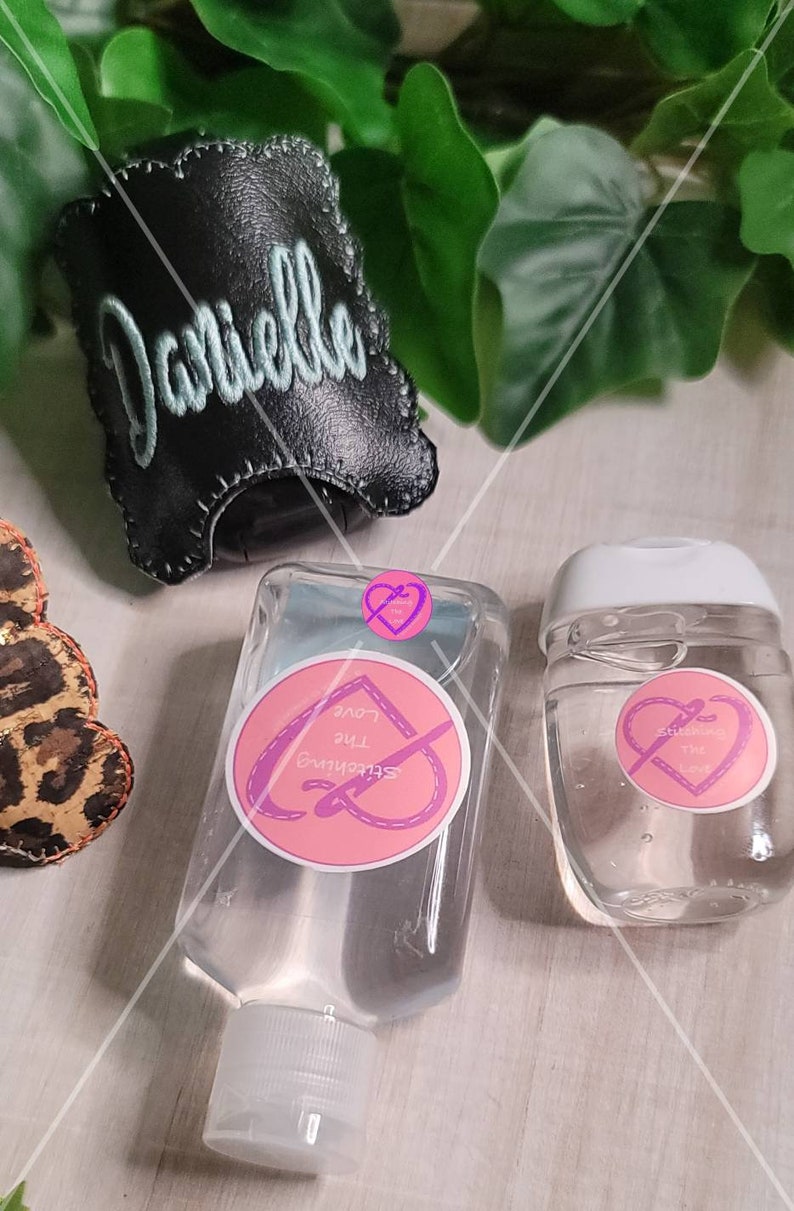 Może przedstawiać: Dwie przezroczyste plastikowe butelki z płynem do dezynfekcji rąk z r&oacute;żowymi i białymi etykietami z napisem "Stitching the Love". Większa butelka ma czarną sk&oacute;rzaną obudowę z wyhaftowanym imieniem "Danielle".