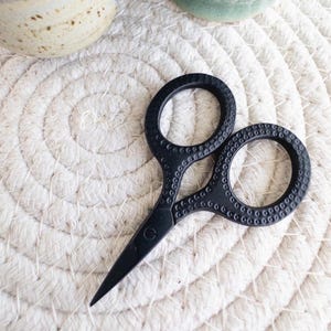 May include: Black embroidery scissors with detailed handle design, resting on a white rope-textured surface. A small ceramic cup with needles and a green mug are in the background. The scissors are a crafting tool.