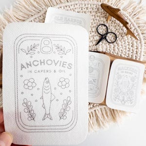 A hand holds a stick and stitch embroidery pattern designed to look like a tin can labeled "Anchovies in Capers & Oil," featuring a line drawing of a fish surrounded by floral and leafy elements. The pattern is placed above a round woven mat with fringe, which also holds additional embroidery patterns with similar tinned fish designs, a pair of black embroidery scissors, and a wooden fish figure as a prop.