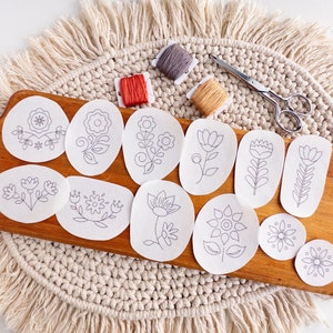 May include: A set of 11 embroidery patterns on white paper, featuring various floral designs. The patterns are arranged on a wooden board with a crocheted surface underneath. The patterns are ready for embroidery projects.