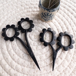 May include: Black flower-shaped scissors and a small ceramic cup with needles on a white woven mat. The scissors have a unique floral design and are likely used for crafting or sewing. The cup is blue and white.