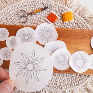 May include: A set of white embroidery patterns featuring different sun designs. The patterns are on a wooden surface with a pair of scissors and two spools of thread.