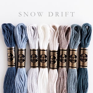 Eight skeins of DMC Mouliné Special embroidery thread in assorted colors: medium navy, light navy, silver, snow white, off-white, gray-brown, medium aqua, dark aqua. Each skein is labeled with DMC branding and specifications. The text 'SNOW DRIFT' is displayed above the threads.