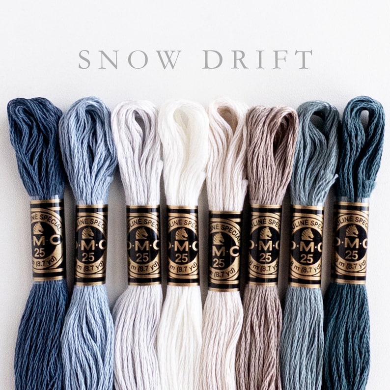 Eight skeins of DMC Mouliné Special embroidery thread in assorted colors: medium navy, light navy, silver, snow white, off-white, gray-brown, medium aqua, dark aqua. Each skein is labeled with DMC branding and specifications. The text 'SNOW DRIFT' is displayed above the threads.