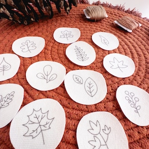 May include: A set of 10 white embroidery hoops with different leaf and floral designs. The hoops are arranged on a brown and orange textured surface.