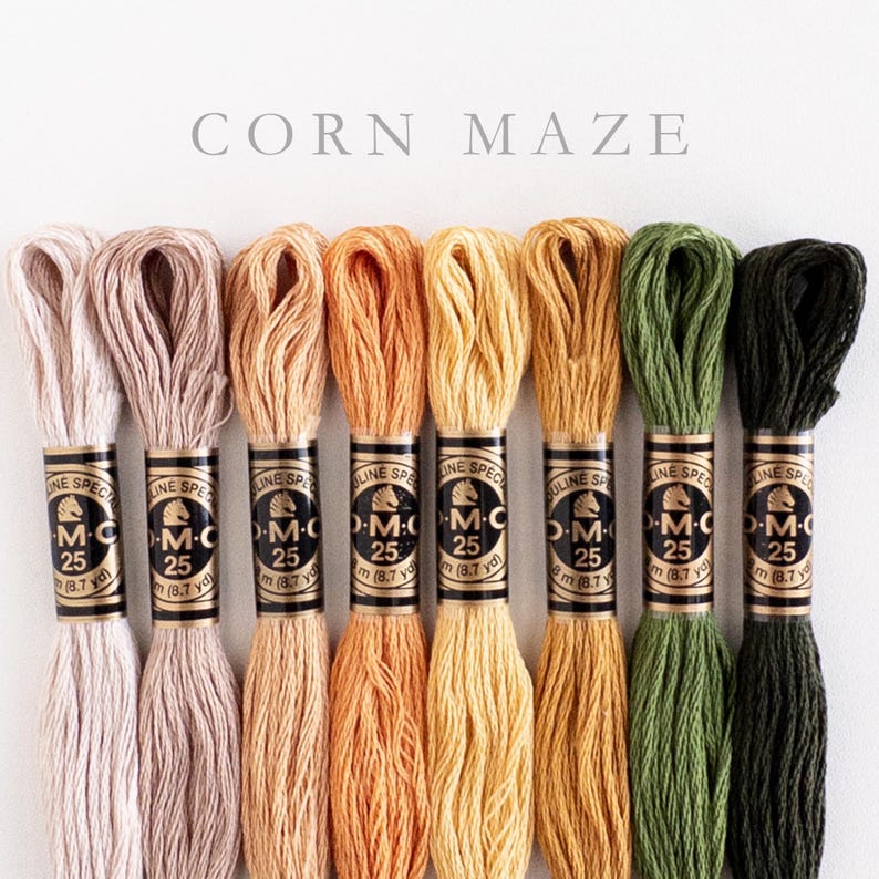 Eight skeins of DMC Mouliné Special embroidery thread in assorted colors: cream, beige, light orange, medium orange, light yellow, gold, forest green, dark forest green. Each skein is labeled with DMC branding and specifications. The text 'CORN MAZE' is displayed above the threads.