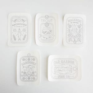 Five stick and stitch embroidery patterns are displayed on a white surface. Each features a hand-drawn tinned fish label: Anchovies in capers and oil, Trout in lemon dill sauce, Mackerel in brine, Sardines in olive oil, and Old Harbor Fish Co. All designs include fish illustrations with floral or decorative elements.