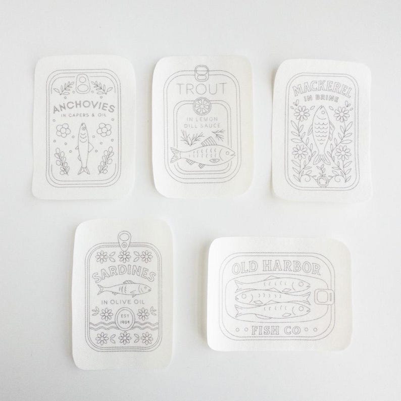 Five stick and stitch embroidery patterns are displayed on a white surface. Each features a hand-drawn tinned fish label: Anchovies in capers and oil, Trout in lemon dill sauce, Mackerel in brine, Sardines in olive oil, and Old Harbor Fish Co. All designs include fish illustrations with floral or decorative elements.