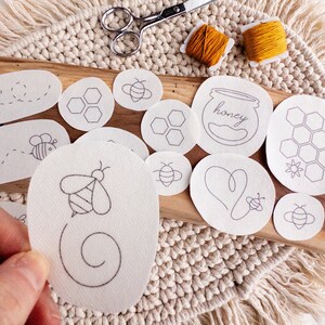 Close-up of a bee-themed stick and stitch embroidery pattern being held by a person. In the background are various designs, including a beehive, a honey jar, a heart, a bee, and a wreath.  The patterns are arranged on a wooden surface with a woven mat background.