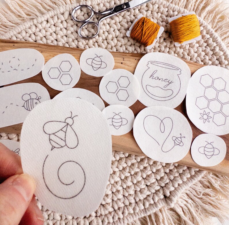 Close-up of a bee-themed stick and stitch embroidery pattern being held by a person. In the background are various designs, including a beehive, a honey jar, a heart, a bee, and a wreath.  The patterns are arranged on a wooden surface with a woven mat background.
