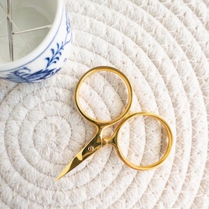 May include: Close-up of gold-colored embroidery scissors with sharp, pointed blades. The scissors are on a white, textured, circular surface. A small, white and blue ceramic container with needles is in the background.