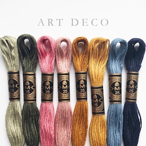 Eight skeins of DMC Mouliné Special embroidery thread in assorted colors: olive green, rose pink, dusty rose, golden yellow, mustard yellow, steel blue, and navy blue. Each skein is labeled with DMC branding and specifications. The text 'ART DECO' is displayed above the threads.