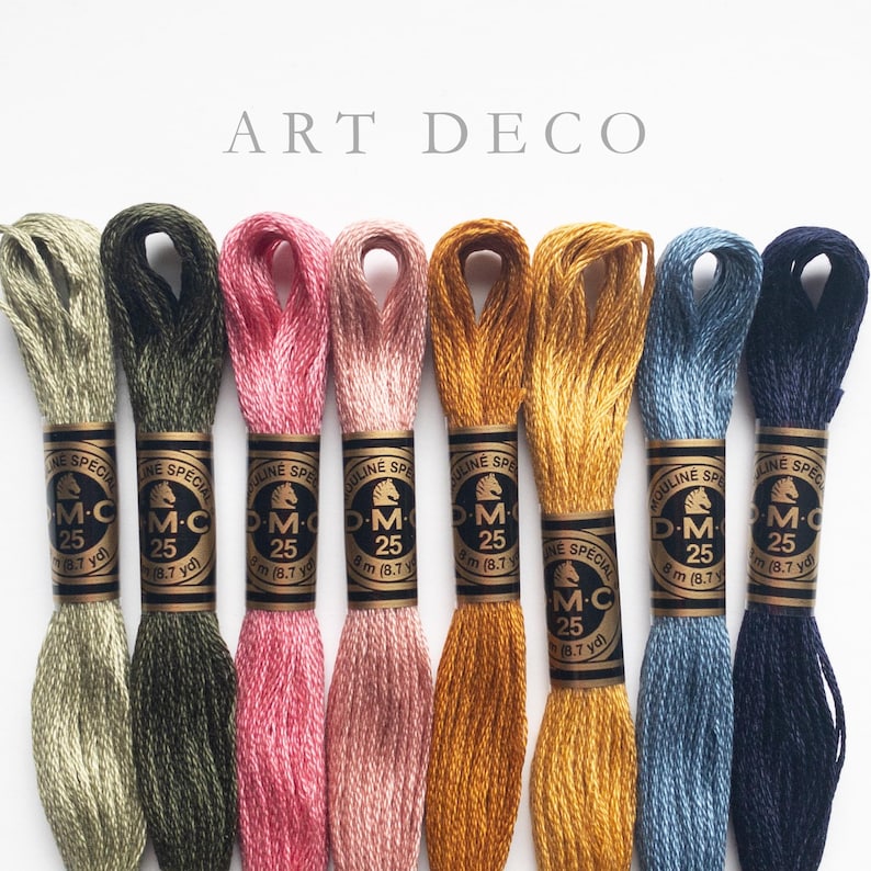 Eight skeins of DMC Mouliné Special embroidery thread in assorted colors: olive green, rose pink, dusty rose, golden yellow, mustard yellow, steel blue, and navy blue. Each skein is labeled with DMC branding and specifications. The text 'ART DECO' is displayed above the threads.