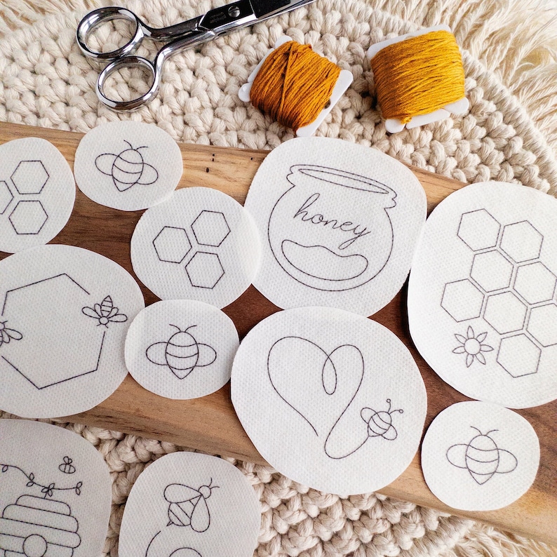 White embroidery stickers with black designs of bees, honeycombs, and a jar of honey that says 'honey' on it. The patterns are on a wooden surface with a cream-colored woven background.