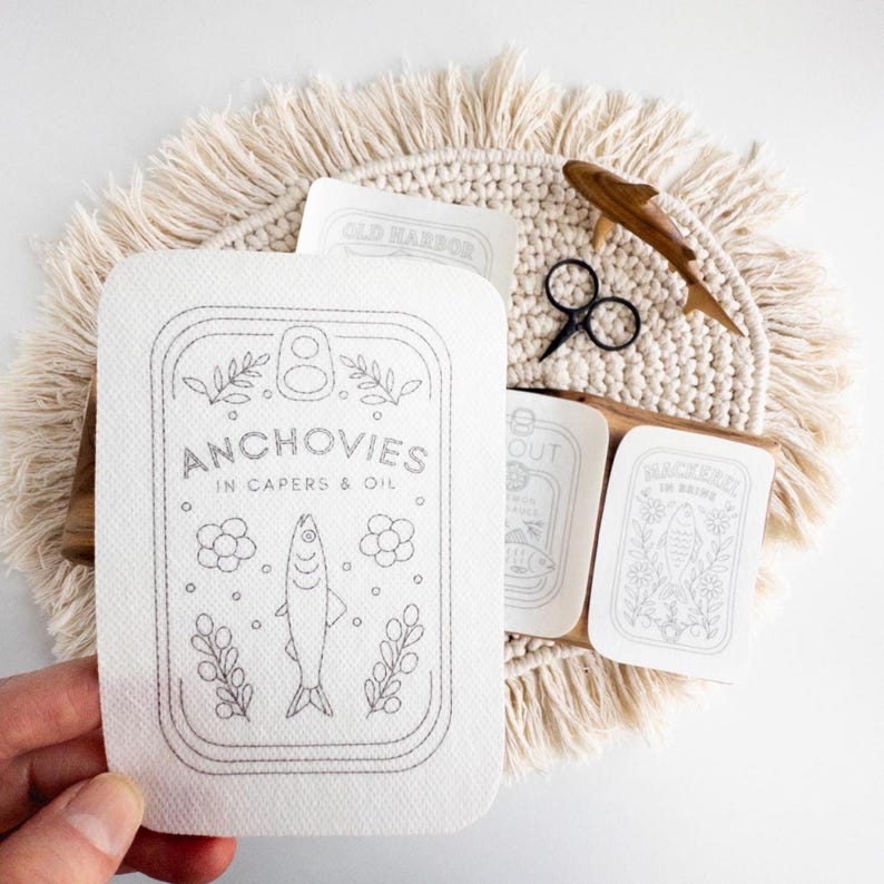 A hand holds a stick and stitch embroidery pattern designed to look like a tin can labeled "Anchovies in Capers & Oil," featuring a line drawing of a fish surrounded by floral and leafy elements. The pattern is placed above a round woven mat with fringe, which also holds additional embroidery patterns with similar tinned fish designs, a pair of black embroidery scissors, and a wooden fish figure as a prop.