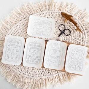 Five stick and stitch embroidery patterns are displayed on a round, fringed woven mat. Designs include: “Old Harbor Fish Co.” with two fish in a tin, “Anchovies in Capers & Oil,” “Sardines in Olive Oil,” “Trout in Lemon Dill Sauce,” and “Mackerel in Brine.” A pair of black embroidery scissors and a wooden fish-shaped thread holder rest nearby on the mat.