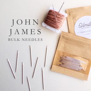 May include: A close-up of a package of 25 embroidery needles, size 5, from John James Bulk Needles. The needles are silver and are packaged in a clear plastic bag with a brown paper backing. The package is labeled "Olmsted Needlework" and has a white circle with the company name in black text.