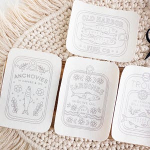 Three stick and stitch embroidery patterns are displayed on a woven mat and wooden surface. Designs include: “Old Harbor Fish Co.” with two fish in a tin, “Trout in Lemon Dill Sauce” with herbs and a lemon slice, and “Mackerel in Brine” surrounded by flowers and leaves. A small pair of black embroidery scissors and a wooden thread holder shaped like a fish are also visible.