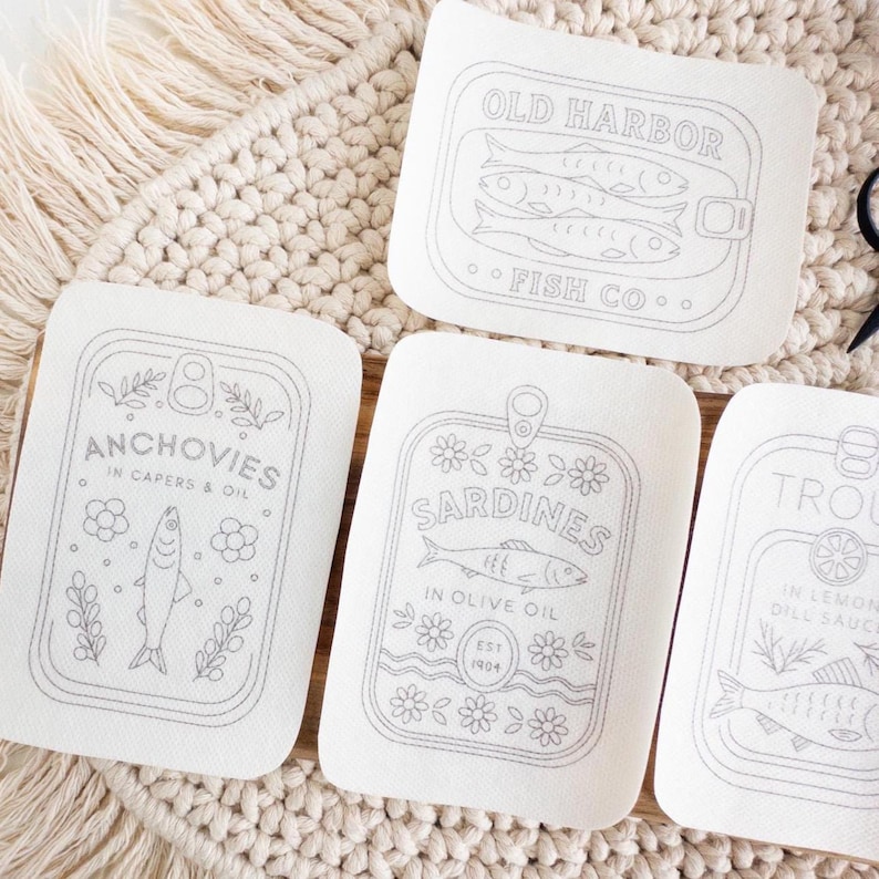 Three stick and stitch embroidery patterns are displayed on a woven mat and wooden surface. Designs include: “Old Harbor Fish Co.” with two fish in a tin, “Trout in Lemon Dill Sauce” with herbs and a lemon slice, and “Mackerel in Brine” surrounded by flowers and leaves. A small pair of black embroidery scissors and a wooden thread holder shaped like a fish are also visible.
