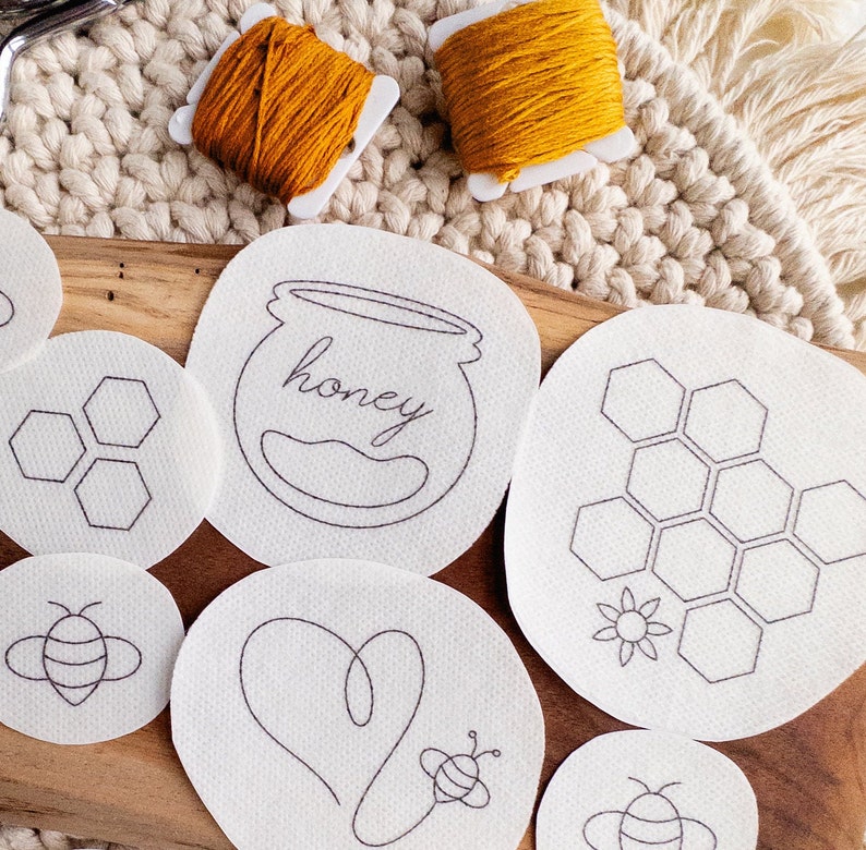 Close-up of dissolvable embroidery designs on a cream-colored woven surface. The patterns include bees, honeycombs, and hearts. The patterns are arranged next to pair of scissors and two bobbins of thread. The text 'honey' is visible on one of the patterns.