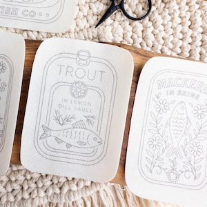 Four stick and stitch embroidery patterns are arranged on a woven mat and wooden surface. Designs include “Old Harbor Fish Co.” with two fish in a tin, “Sardines in Olive Oil” with flowers, “Trout in Lemon Dill Sauce” with herbs and a lemon slice, and “Mackerel in Brine” surrounded by floral elements. A pair of black embroidery scissors and a wooden fish-shaped figure are also visible.