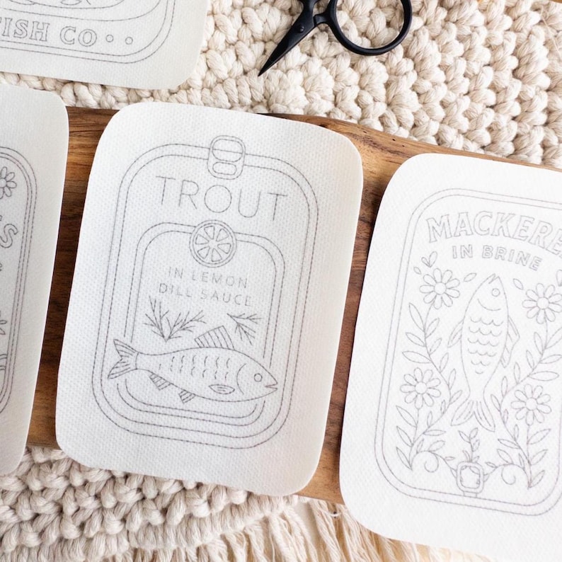 Four stick and stitch embroidery patterns are arranged on a woven mat and wooden surface. Designs include “Old Harbor Fish Co.” with two fish in a tin, “Sardines in Olive Oil” with flowers, “Trout in Lemon Dill Sauce” with herbs and a lemon slice, and “Mackerel in Brine” surrounded by floral elements. A pair of black embroidery scissors and a wooden fish-shaped figure are also visible.