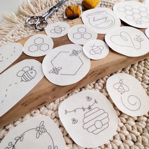 A set of white embroidery pattern transfers on a cream-colored woven surface. The patterns include bees, honeycombs, and hearts. The patterns are arranged next to pair of scissors and two bobbins of thread. The text 'honey' is visible on one of the patterns.