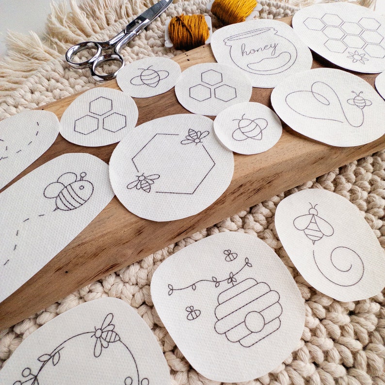 A set of white embroidery pattern transfers on a cream-colored woven surface. The patterns include bees, honeycombs, and hearts. The patterns are arranged next to pair of scissors and two bobbins of thread. The text 'honey' is visible on one of the patterns.
