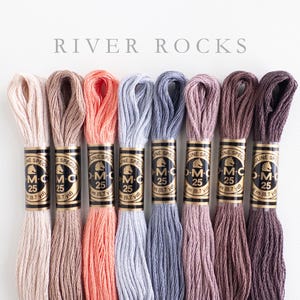 Eight skeins of DMC Mouliné Special embroidery thread in assorted colors: cream, beige, coral, light blue, dusty blue, light cocoa, dark cocoa, extra dark cocoa. Each skein is labeled with DMC branding and specifications. The text 'RIVER ROCKS' is displayed above the threads.