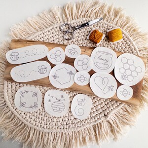 A set of white stick and stitch embroidery patterns on a cream-colored woven surface. The patterns include bees, honeycombs, and hearts. The patterns are arranged next to pair of scissors and two bobbins of thread. The text 'honey' is visible on one of the patterns.