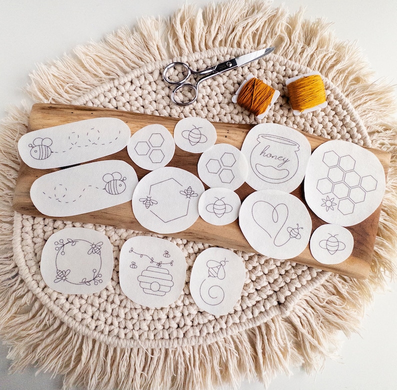A set of white stick and stitch embroidery patterns on a cream-colored woven surface. The patterns include bees, honeycombs, and hearts. The patterns are arranged next to pair of scissors and two bobbins of thread. The text 'honey' is visible on one of the patterns.