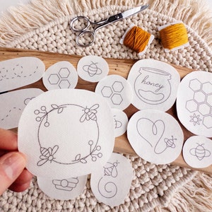 Close-up of a bee-themed stick and stitch embroidery pattern being held by a person. In the background are various designs, including a beehive, a honey jar, a heart, a bee, and a wreath.  The patterns are arranged on a wooden surface with a woven mat background.
