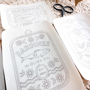 Three stick and stitch embroidery patterns rest on a woven mat and wooden surface. Designs include “Sardines in Olive Oil” with flowers and waves, “Old Harbor Fish Co.” with two fish in a tin, and “Trout in Lemon Dill Sauce” featuring a fish, lemon slice, and herbs. Black embroidery scissors and a wooden fish-shaped thread holder are also visible.