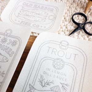 Close-up of three stick and stitch embroidery patterns on a woven mat and wooden surface. Visible designs include “Trout in Lemon Dill Sauce” with a fish, lemon slice, and herbs; “Old Harbor Fish Co.” with two fish in a tin; and part of a “Sardines in Olive Oil” label with floral accents. Black embroidery scissors are also shown.