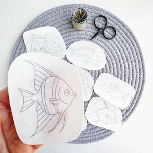 May include: A collection of fish embroidery patterns on white fabric, with a variety of fish designs in black thread. A small pair of black scissors and a small ceramic container with needles are also present. The items are on a gray woven mat.