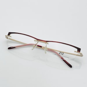 May include: Rimless eyeglasses with a burgundy top frame and temples, and a gold-colored bridge. The lenses are clear, and the glasses have the text "212 CAROLINA HERRERA" on the temple.
