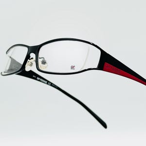 Op de afbeelding: Zwarte bril met een rechthoekig montuur en heldere glazen. De veren zijn zwart met een rode accentkleur en de merknaam "GF Ferre" is zichtbaar. De bril staat tegen een witte achtergrond.