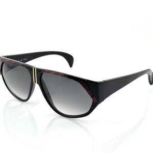 May include: Black sunglasses with a geometric design. The frames have a dark tortoiseshell pattern on the top and sides, with a gold-colored vertical stripe on the front. The lenses are gradient gray.