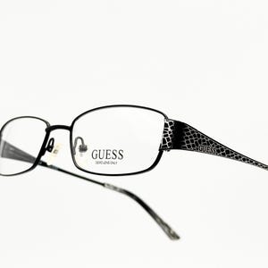 May include: Black rectangular eyeglasses with a black and silver patterned arm. The glasses have the word "GUESS" printed on the frame. "DEMO LENS ONLY" is printed below the word "GUESS".