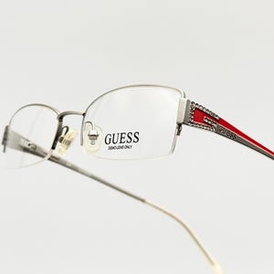 以下が含まれることがあります： シルバーカラーのGuessメガネ。ハーフリムのデザインで、テンプルには赤とシルバーのアクセントがあり、小さな輝くディテールで装飾されています。レンズはクリアで、Guessのロゴが見えます。