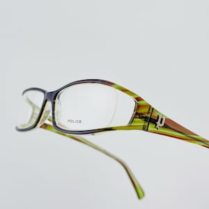 May include: Eyeglasses with a black frame and a green, yellow, and brown patterned design. The lenses are clear, and the word "POLICE" is printed on the frame. The glasses have a unique shape and style.