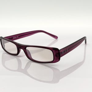 Peut inclure: Lunettes en plastique de couleur violette avec des verres transparents. Les lunettes ont une forme rectangulaire et le nom de la marque "DIXIE" est imprimé sur le côté.