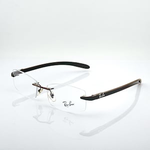 Può includere: Occhiali senza montatura con lenti rettangolari trasparenti e logo Ray-Ban stampato sulla lente destra. La montatura presenta aste sottili color marrone scuro e un ponte metallico con naselli regolabili, caratterizzati da un design leggero.