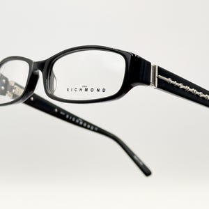 Puede incluir: Gafas rectangulares negras con un acento plateado en las patillas y la palabra "RICHMOND" impresa en la lente derecha.