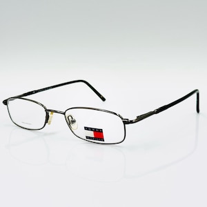 Puede incluir: Gafas rectangulares con montura de metal oscuro. Las lentes son transparentes y las patillas negras. El logotipo de Tommy Hilfiger es visible en la lente derecha. Sobre fondo blanco.