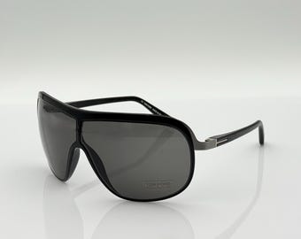 May include: Black sunglasses with a wraparound design. The sunglasses have a thick black frame and dark gray lenses. The temples are black with a silver accent. The brand name "TOM FORD" is visible on the lens.