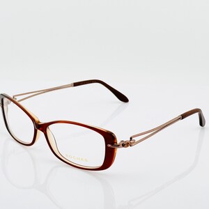 Puede incluir: Gafas marrones y doradas con montura rectangular. Las gafas tienen una montura marrón claro con varillas y bisagras doradas. La marca ROCHAS es visible en la lente.