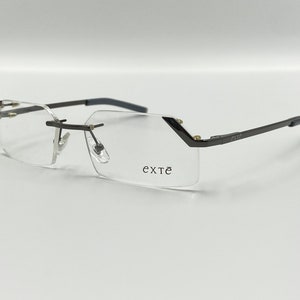 May include: A pair of rimless eyeglasses with a dark gray metal frame. The frame has a subtle angular design and the brand name "exté" is printed on the right lens.