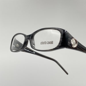 May include: Black and silver eyeglasses with a rectangular frame. The brand name "roberto cavalli" is printed on the right lens.